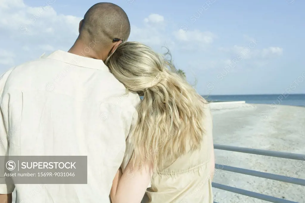 Couple standing arm in arm looking at view, woman's head on man's shoulder, rear view