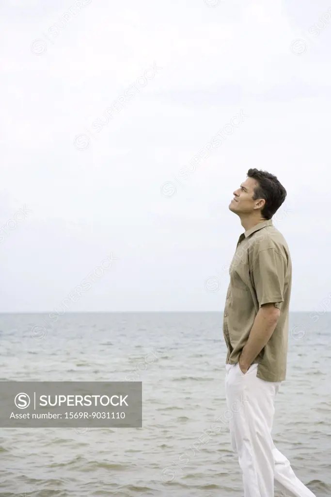 Man walking beside ocean with hands in pockets, looking up