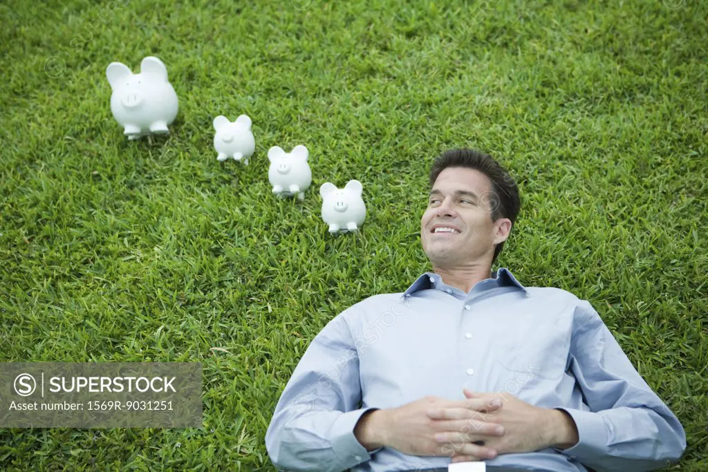 Man lying on grass, next to piggy banks, smiling