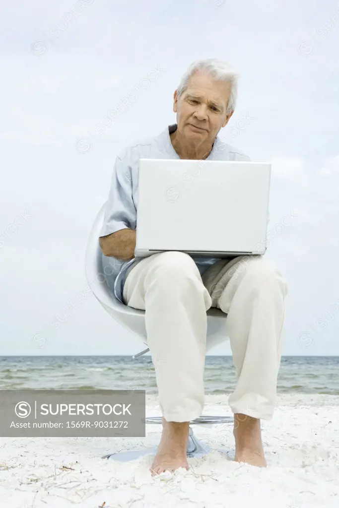 Senior man sitting in chair on beach, using laptop