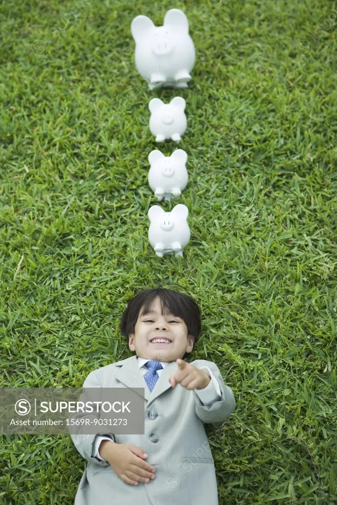 Boy dressed in suit lying on grass, laughing and pointing at camera, piggy banks above head