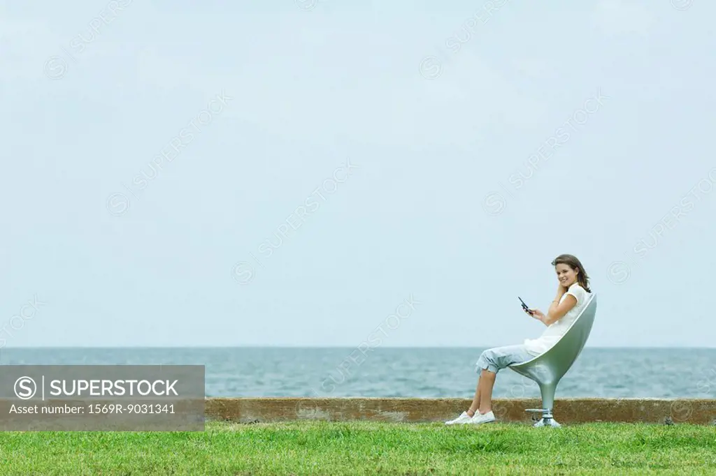 Teenage girl sitting in chair by the sea, holding cell phone