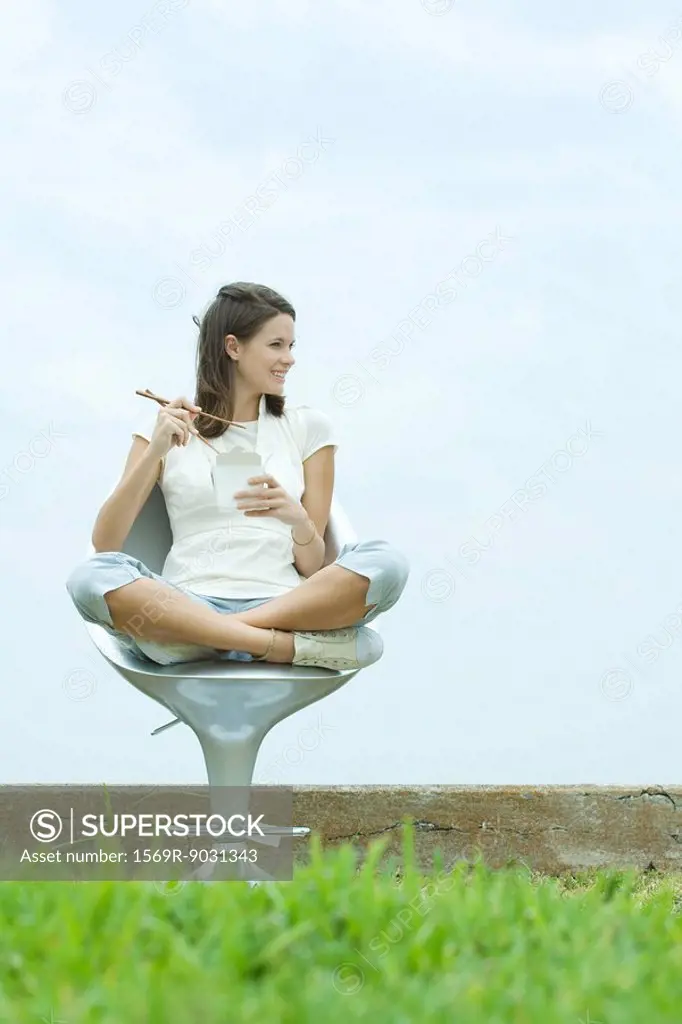 Teenage girl sitting in chair outdoors, holding chopsticks and take out food container, looking away