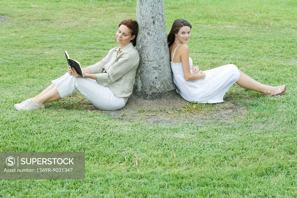 Mother and daughter leaning against tree trunk, woman reading book, teen girl smiling at camera