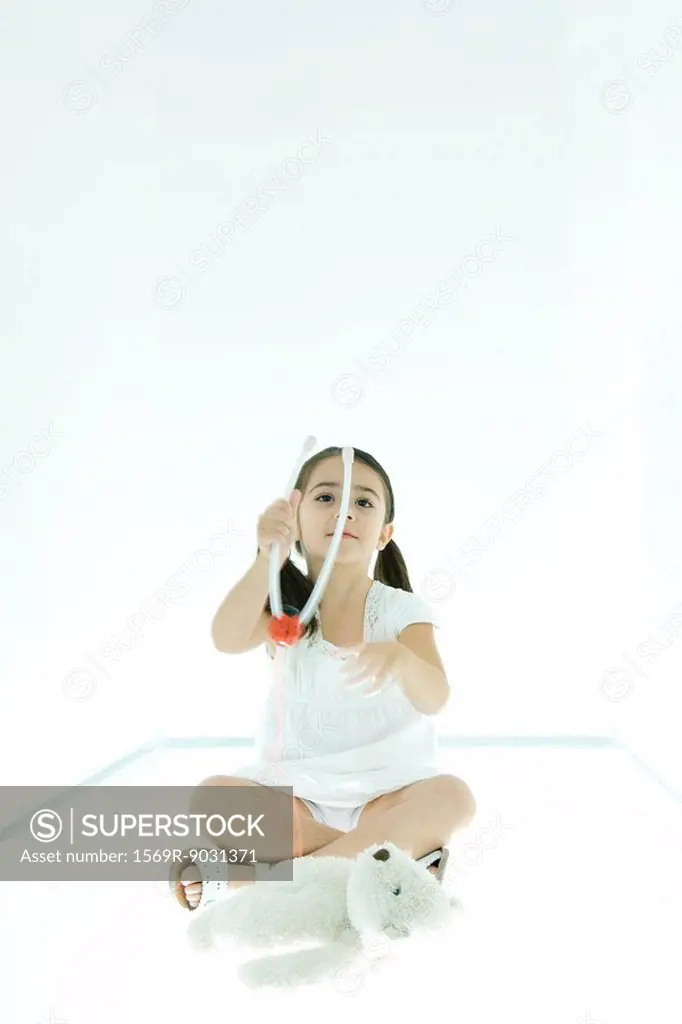 Little girl sitting next to teddy bear, playing with toy stethoscope