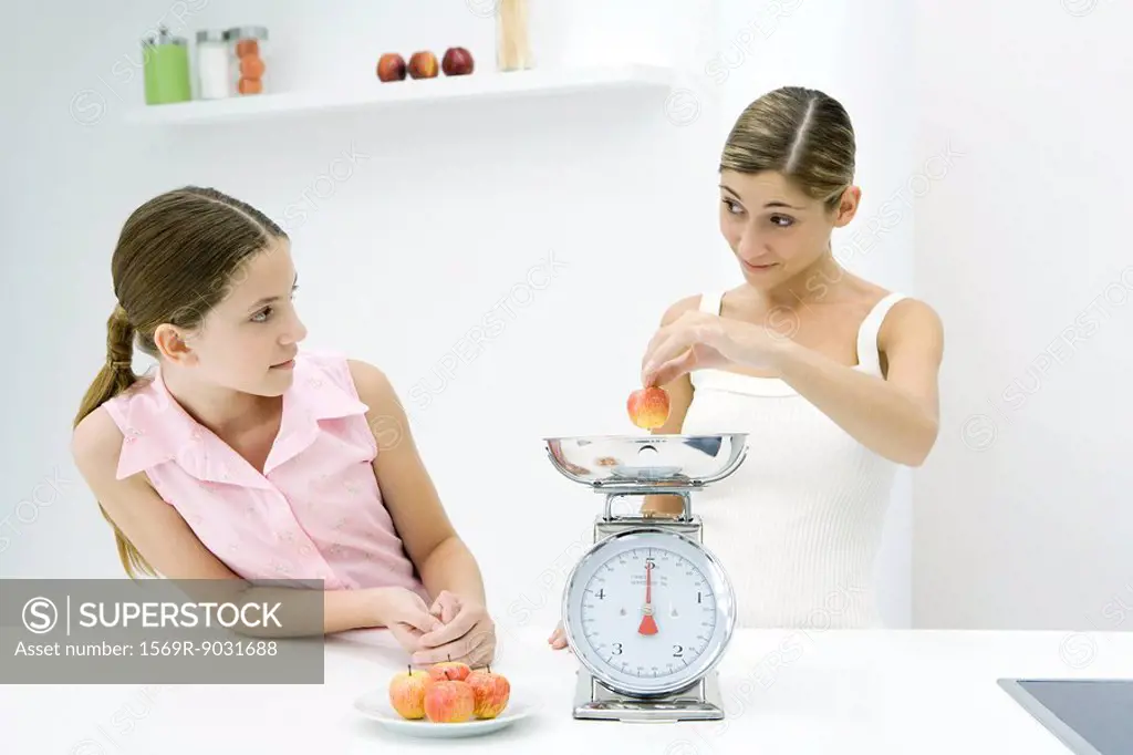 Mother and daughter weighing apples on scale, looking at each other