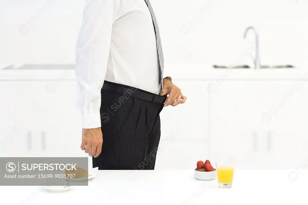 Man standing in kitchen beside two breakfast options, holding waistline of pants, cropped