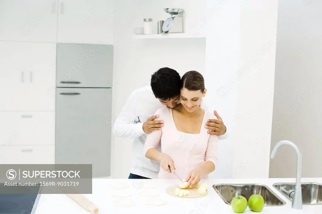 Couple standing together in kitchen, woman cutting apples, man leaning head on her shoulder