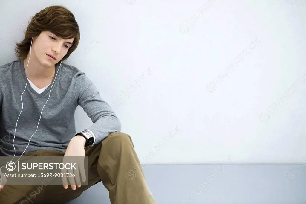 Young man sitting on the ground, listening to earphones, looking away
