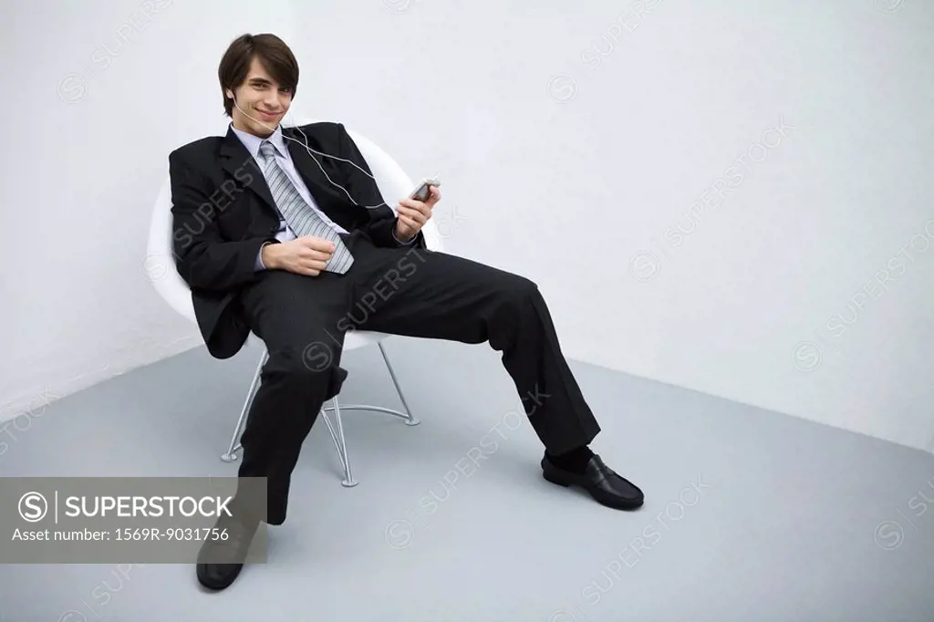 Young man in suit leaning back in chair, listening to mp3 player, smiling at camera, full length