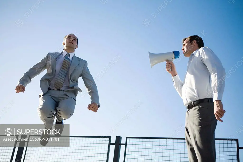 Businessman shouting into megaphone, colleague jumping in the air, low angle view