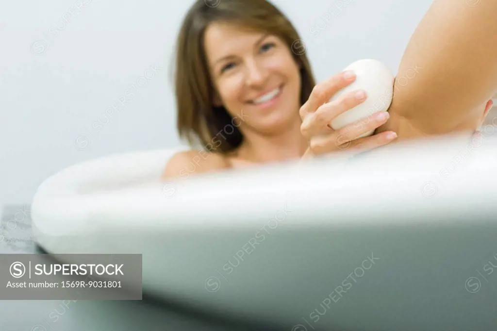 Woman sitting in bathtub, washing leg with soap, smiling