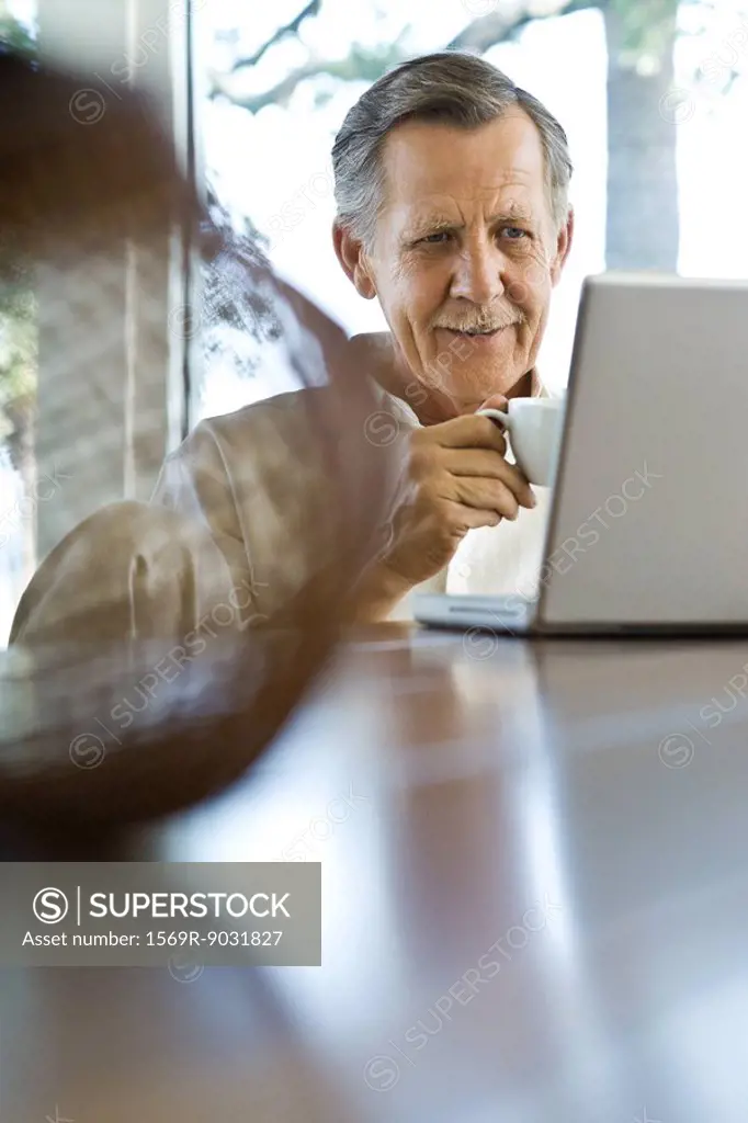 Senior man sitting at table, using laptop computer, holding coffee cup