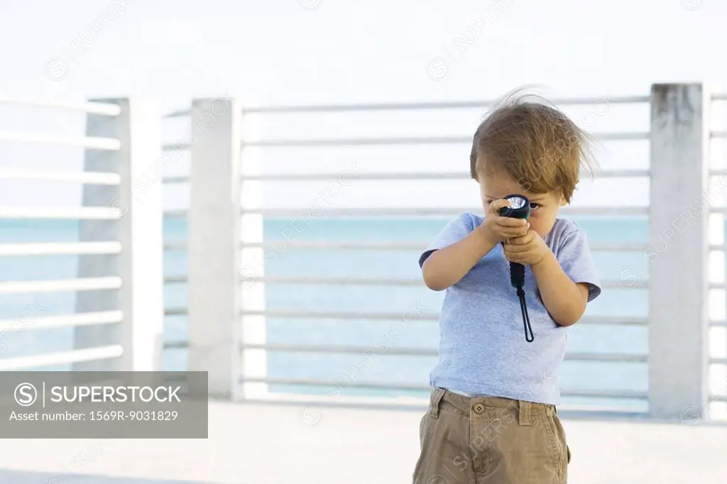 Toddler boy standing outdoors, hiding behind flashlight, peeking at camera