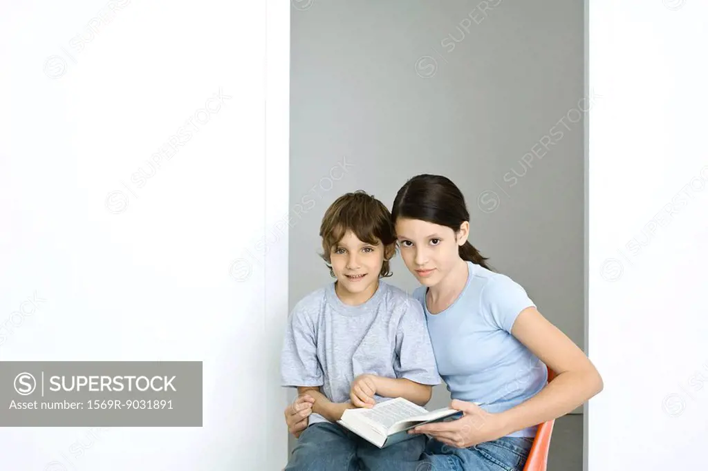 Brother and sister sitting together, girl holding book, both looking at camera