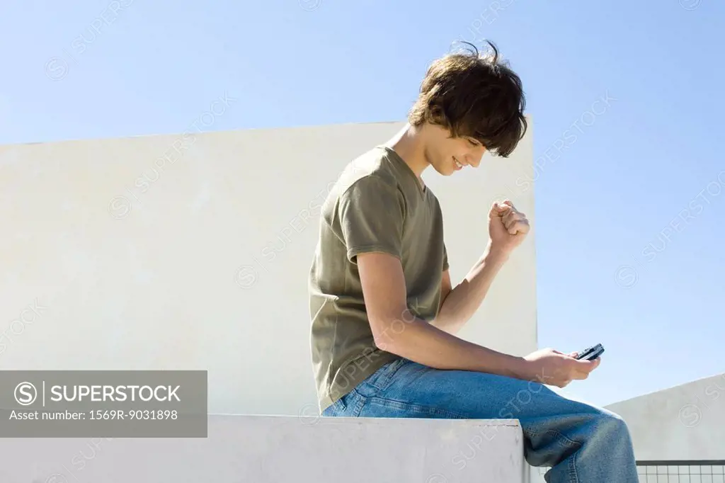 Teenage boy sitting outdoors, looking down at cell phone, making a fist, smiling