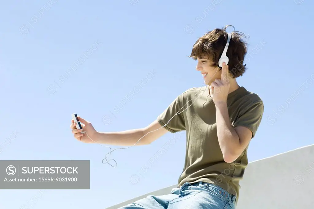 Teenage boy sitting outdoors, listening to headphones, looking at mp3 player, side view