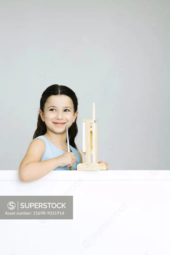 Little girl in front of easel, holding paintbrush, looking away, smiling