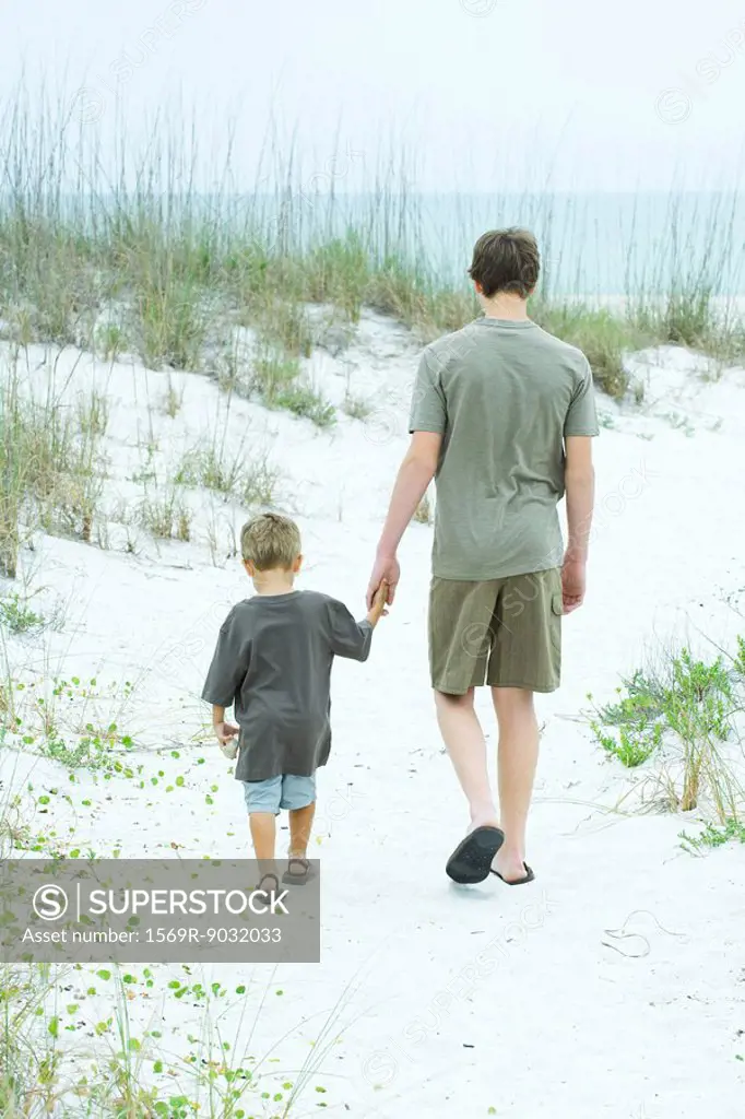 Teenage boy and little brother walking at the beach, holding hands, rear view
