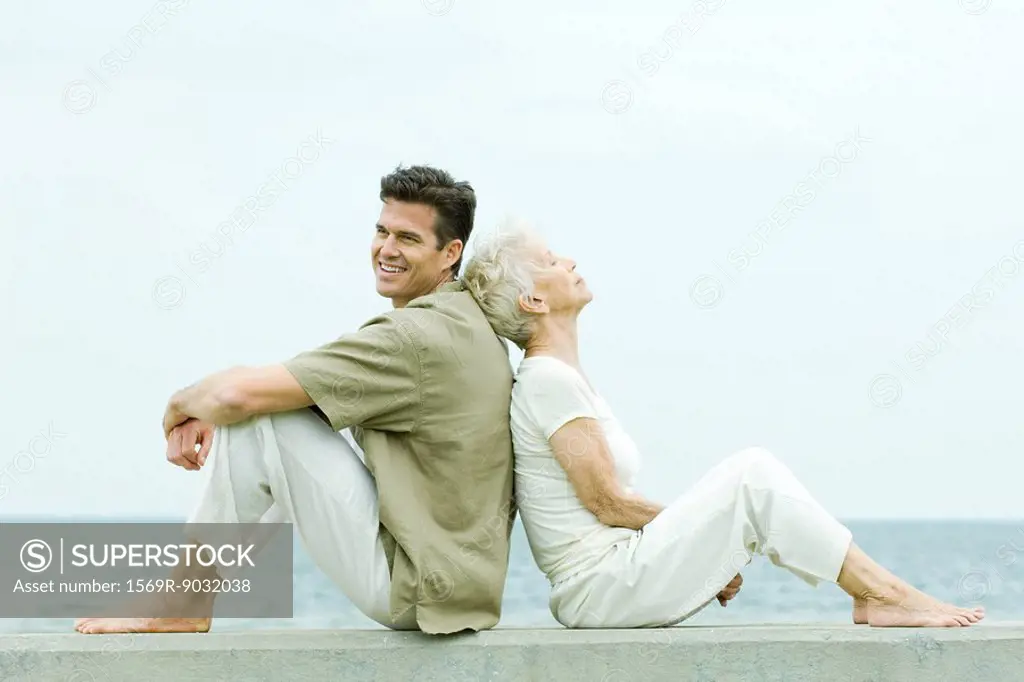 Senior woman and adult son sitting back to back by the sea, side view