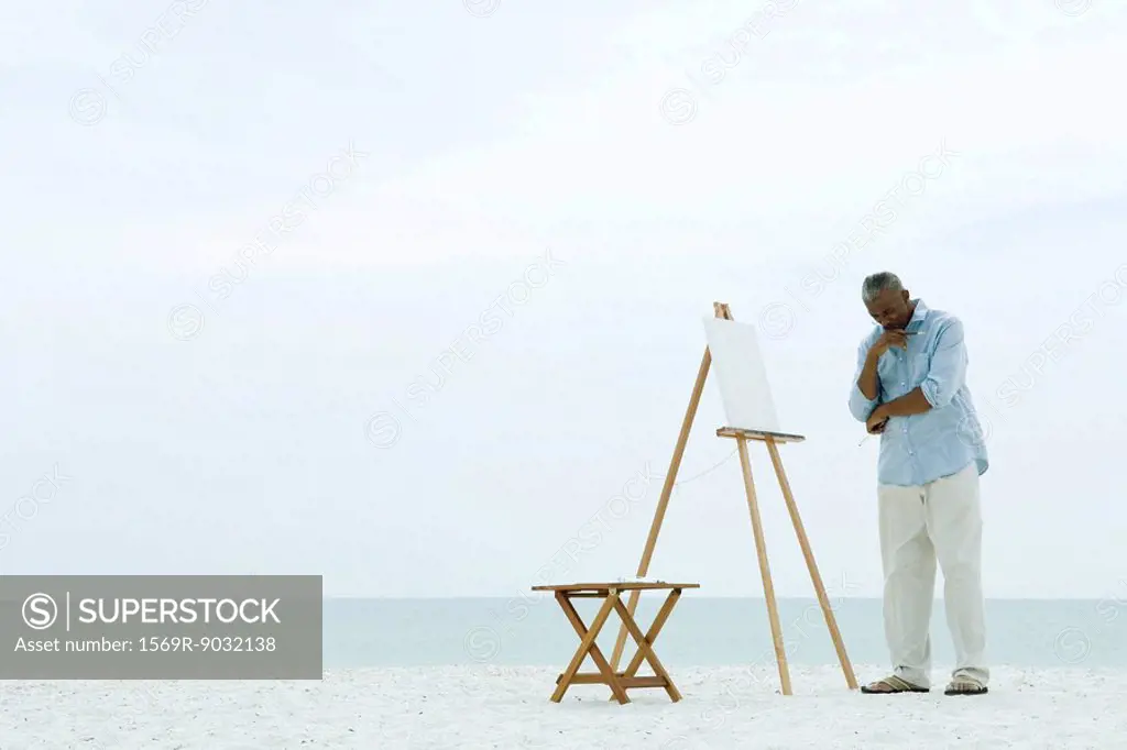 Male painter standing in front of blank canvas at the beach, hand under chin, looking down