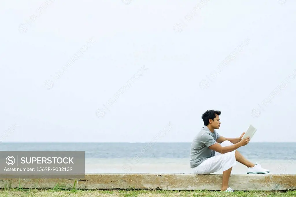 Man sitting on low wall at the beach, reading book, side view
