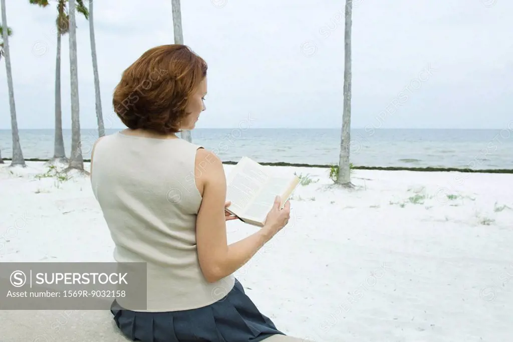 Woman sitting at the beach reading book, rear view