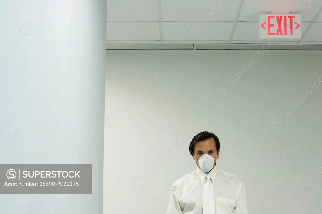 Businessman wearing pollution mask, under exit sign, looking at camera