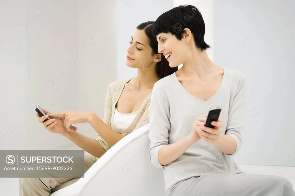 Two women sitting together, both holding cell phones, one looking over shoulder, the other pointing