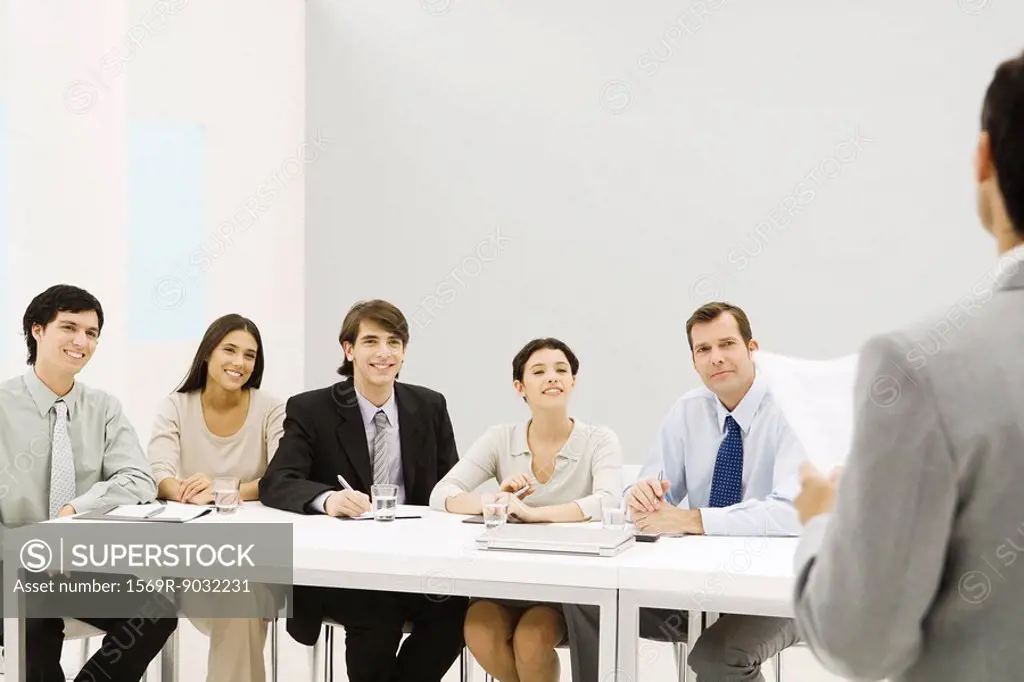 Group of professionals sitting at table, smiling, looking at man standing in foreground