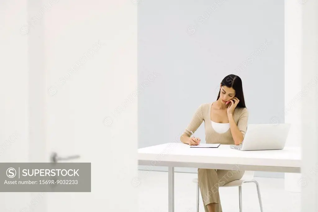 Businesswoman sitting at desk, using cell phone and laptop computer, writing on paper