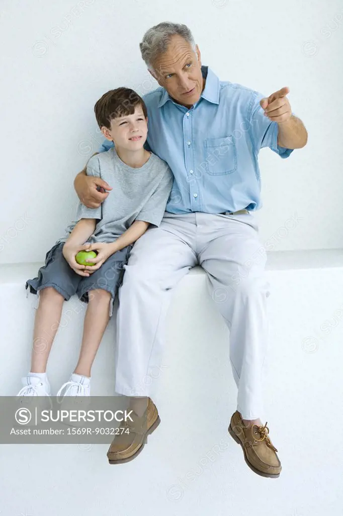 Man sitting with arm around grandson´s shoulders, pointing, both looking away