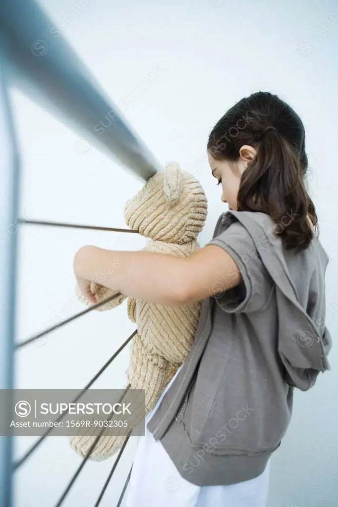 Little girl holding teddy bear, leaning against railing, side view