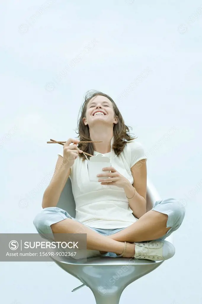 Teenage girl sitting in chair, holding chopsticks and take out food container, eyes closed