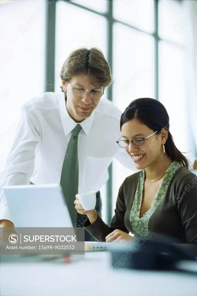 Male and female colleagues looking at laptop computer together, woman holding coffee cup