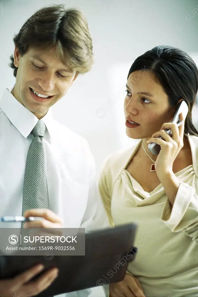 Male and female colleagues standing side by side, woman using cell phone, man looking at binder