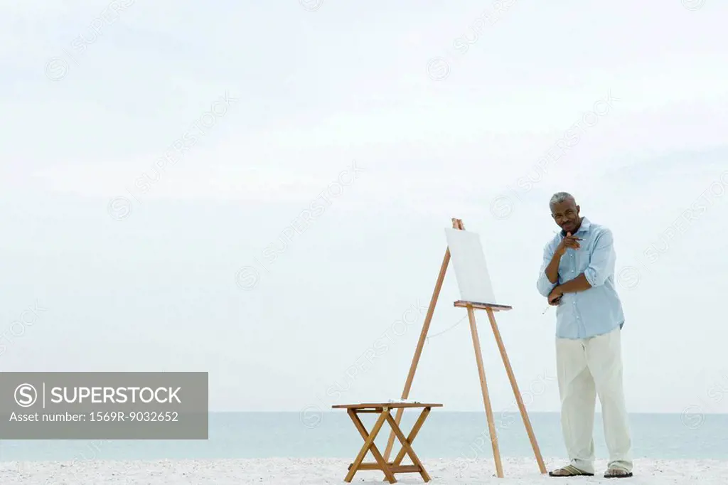 Senior man standing next to blank canvas at the beach, holding paint brushes, smiling at camera