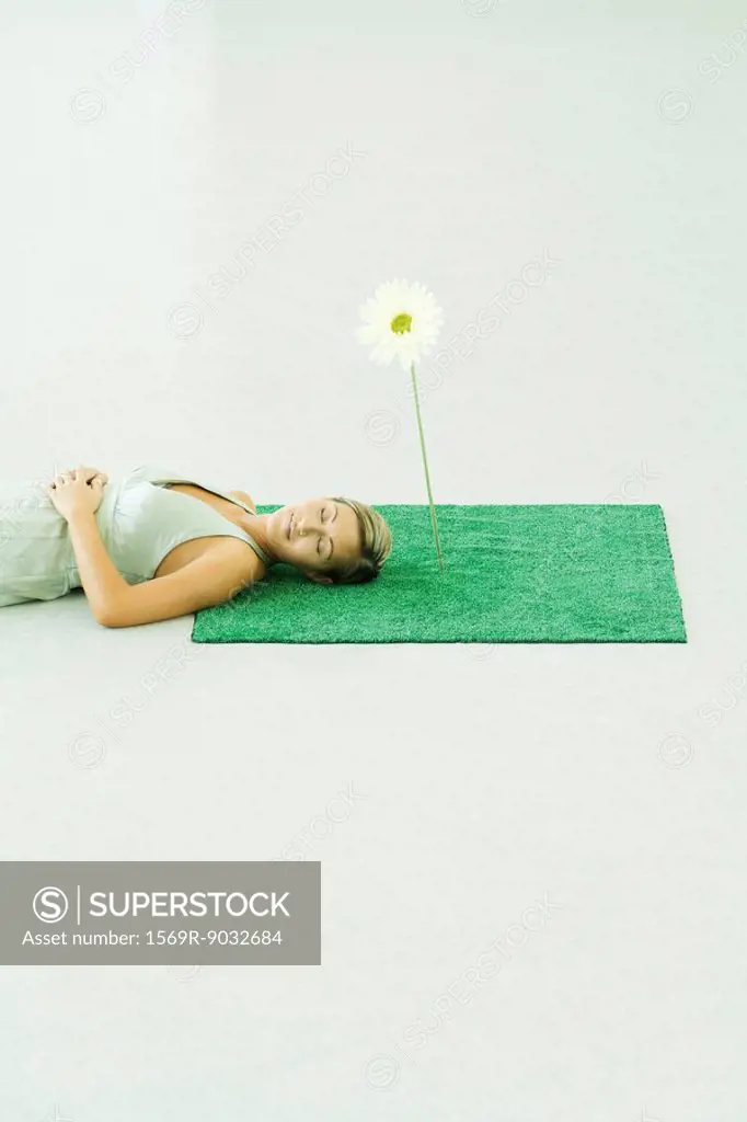 Woman lying on artificial turf next to gerbera daisy, eyes closed