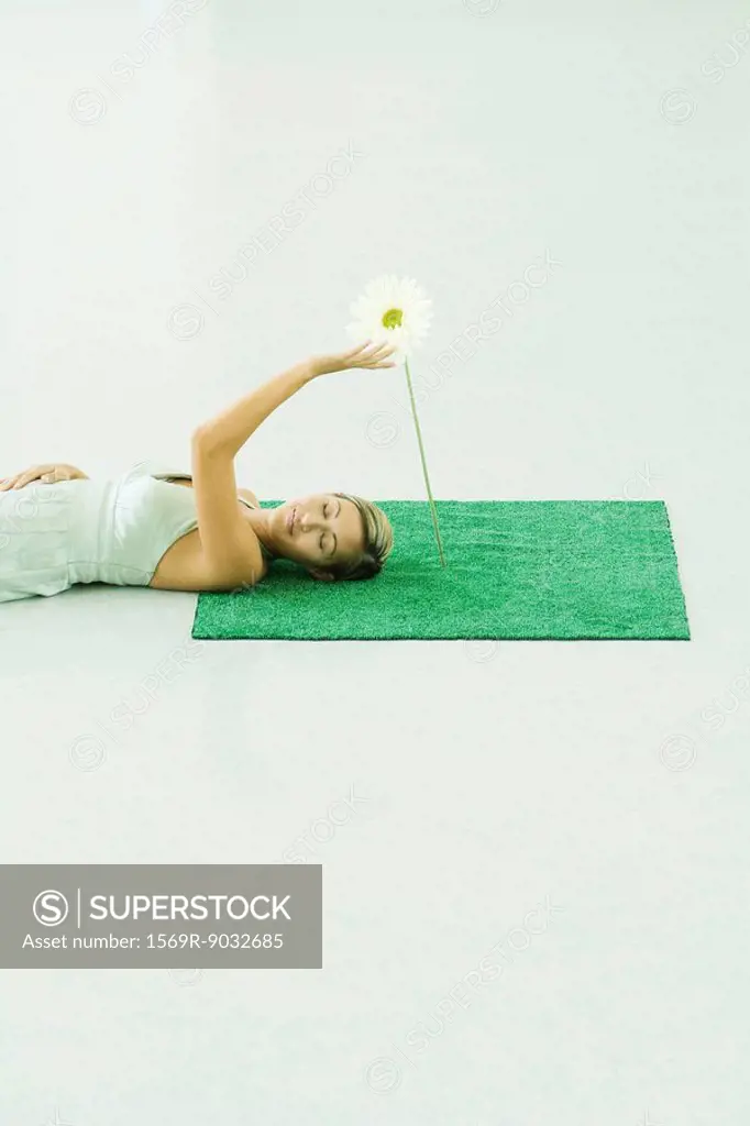 Woman lying on artificial turf, touching gerbera daisy, eyes closed