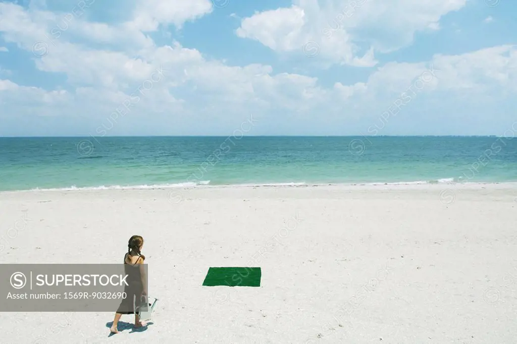 Girl at the beach walking toward patch of artificial turf, carrying watering can, rear view