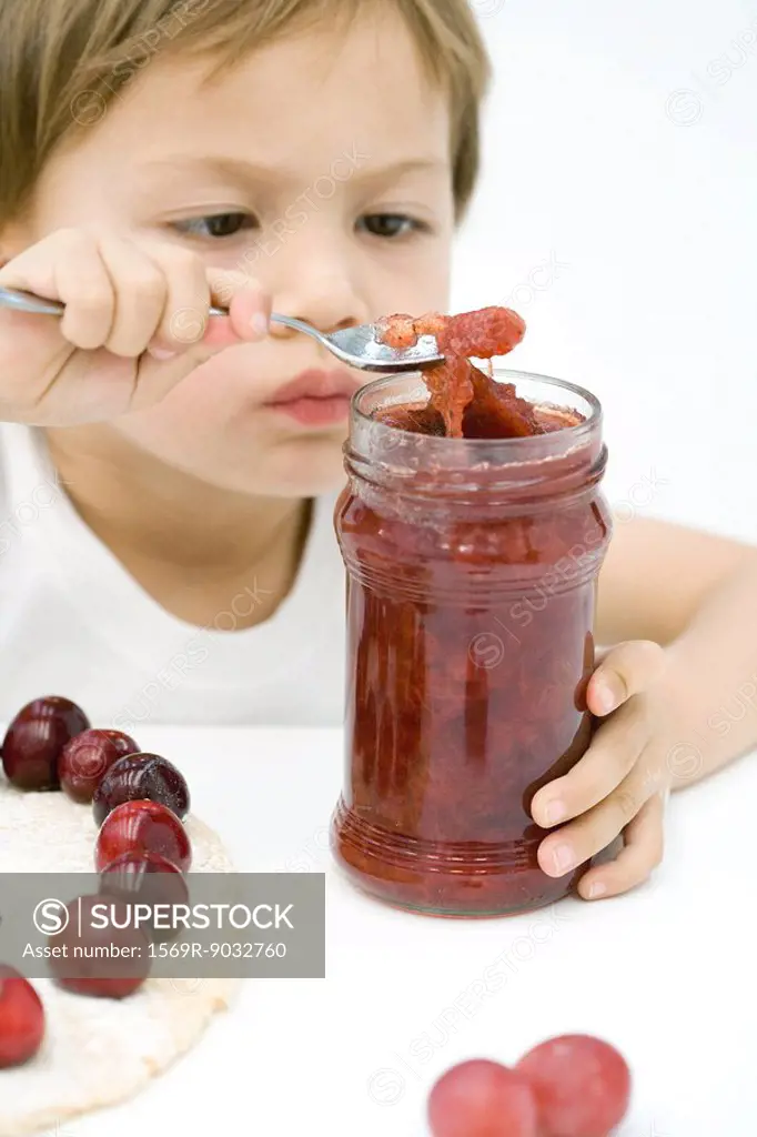 Little boy scooping jam out of jar with spoon, tart and cherries on table in front of him
