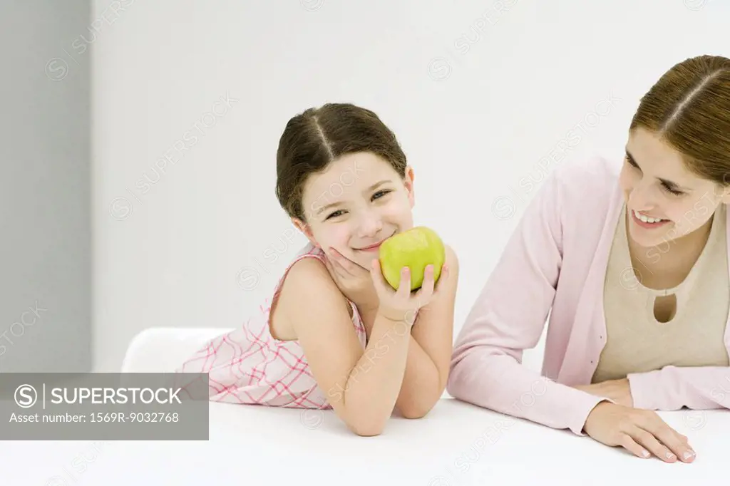 Little girl sitting at table with mother, hand under chin, holding apple, smiling at camera
