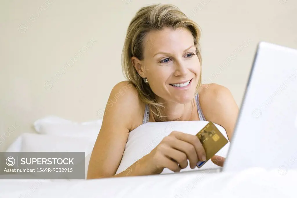 Woman lying in bed, using laptop computer, holding credit card, smiling