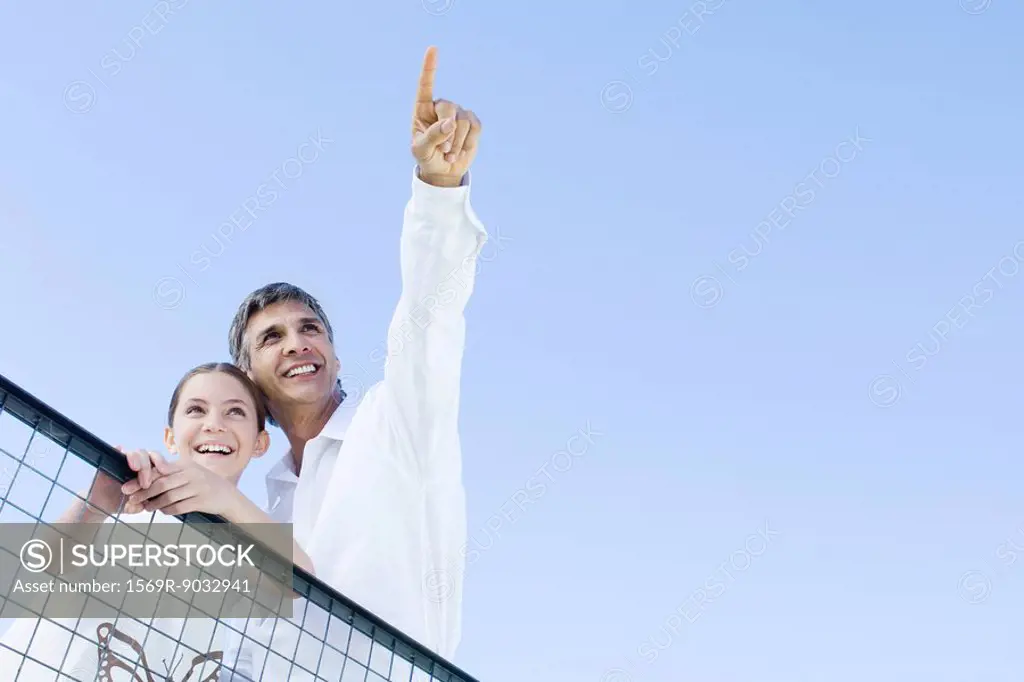 Man standing outdoors with daughter, pointing at the sky, both looking up and smiling, low angle view