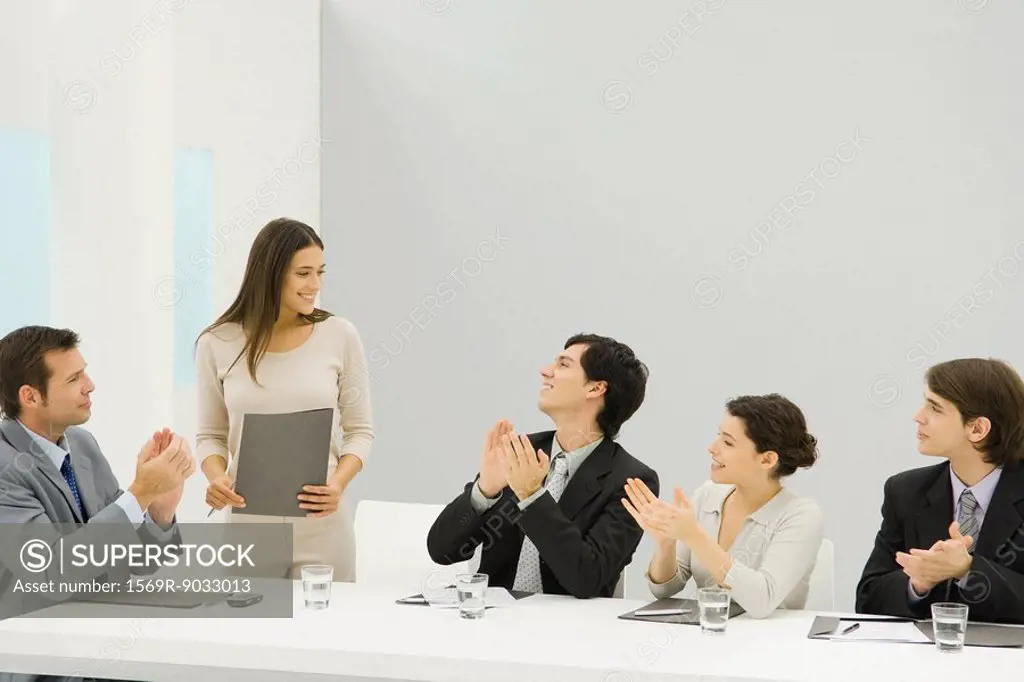 Businesswoman standing beside conference table, holding document, colleagues looking at her and clapping