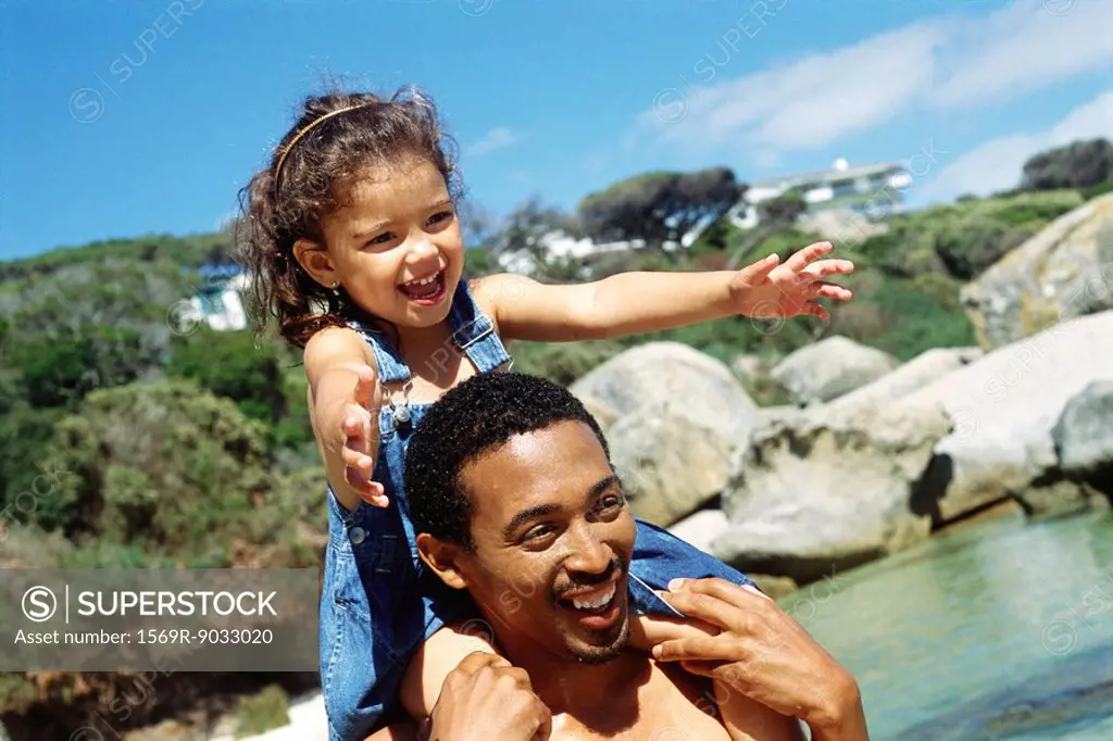 Father carrying daughter on shoulders at the beach, both smiling, close-up