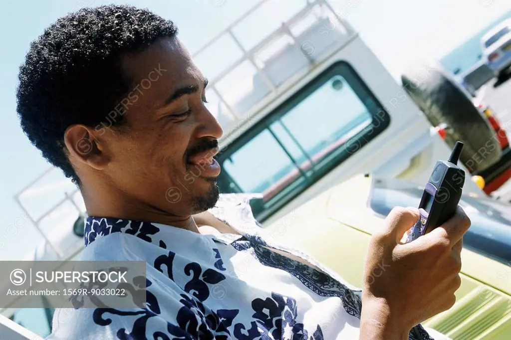 Man in Hawaiian shirt outdoors, looking down at cell phone, smiling