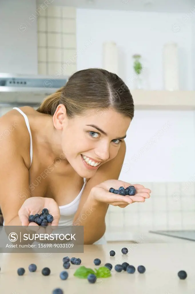 Woman in kitchen, holding handfuls of blueberries, smiling at camera