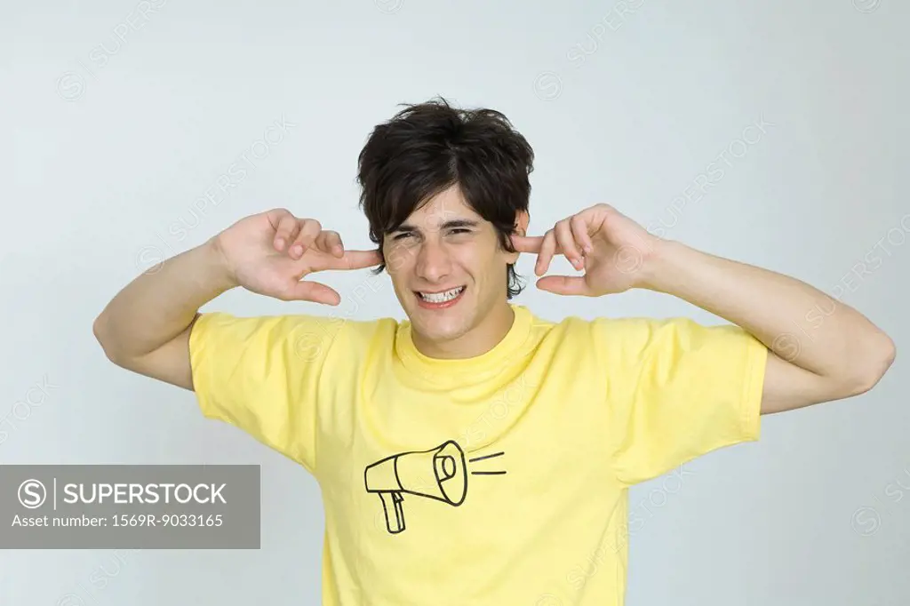 Young man wearing tee-shirt with megaphone graphic, covering ears, looking at camera