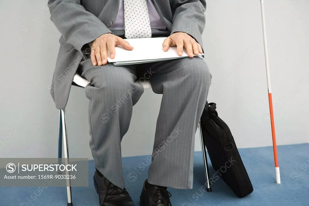 Visually impaired man sitting in chair, holding documents on lap, cropped view, chest down